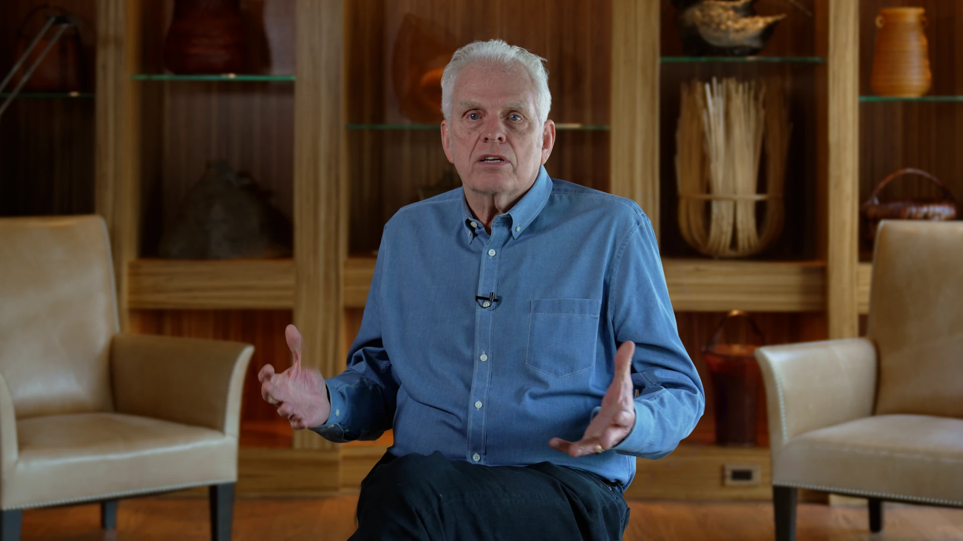 Older man with white hair in a blue button-down shirt speaks, hands raised, in a wood-paneled interview room behind him.