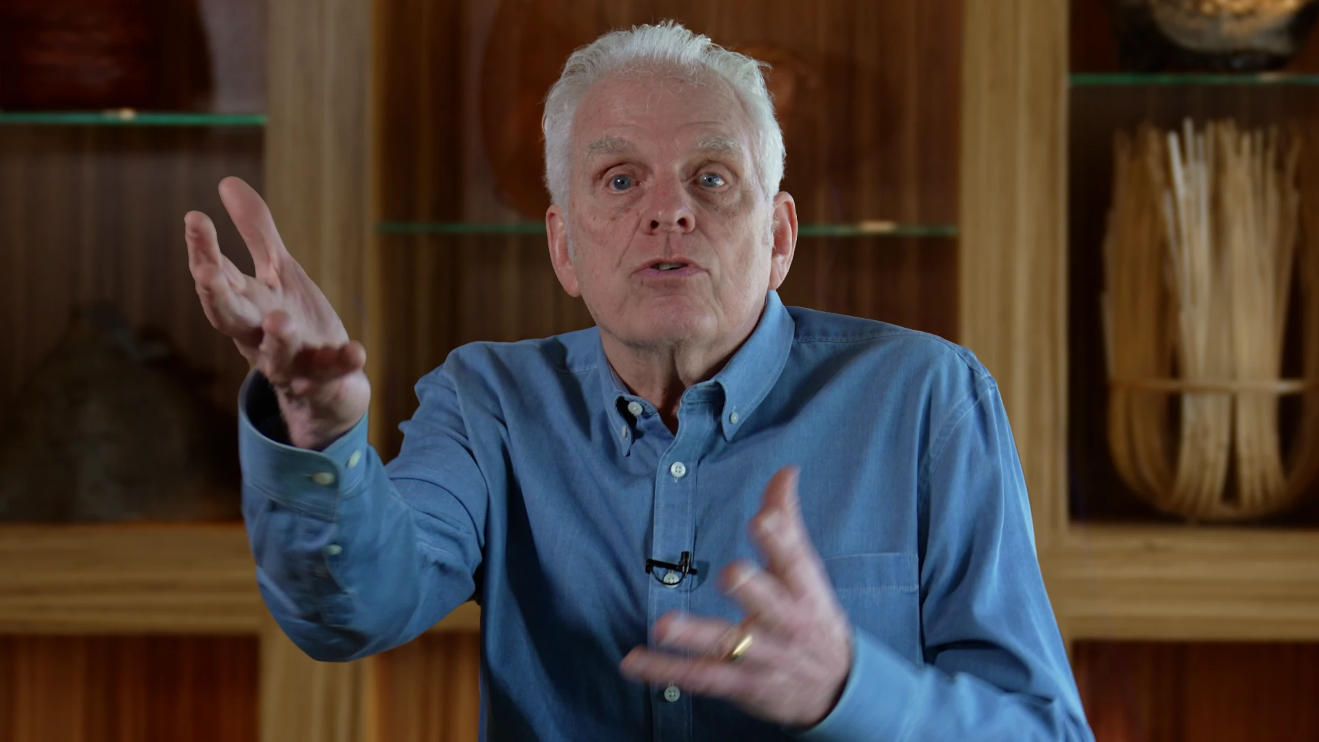 Older man in a blue shirt speaks animatedly, gesturing with hands during a discussion indoors.