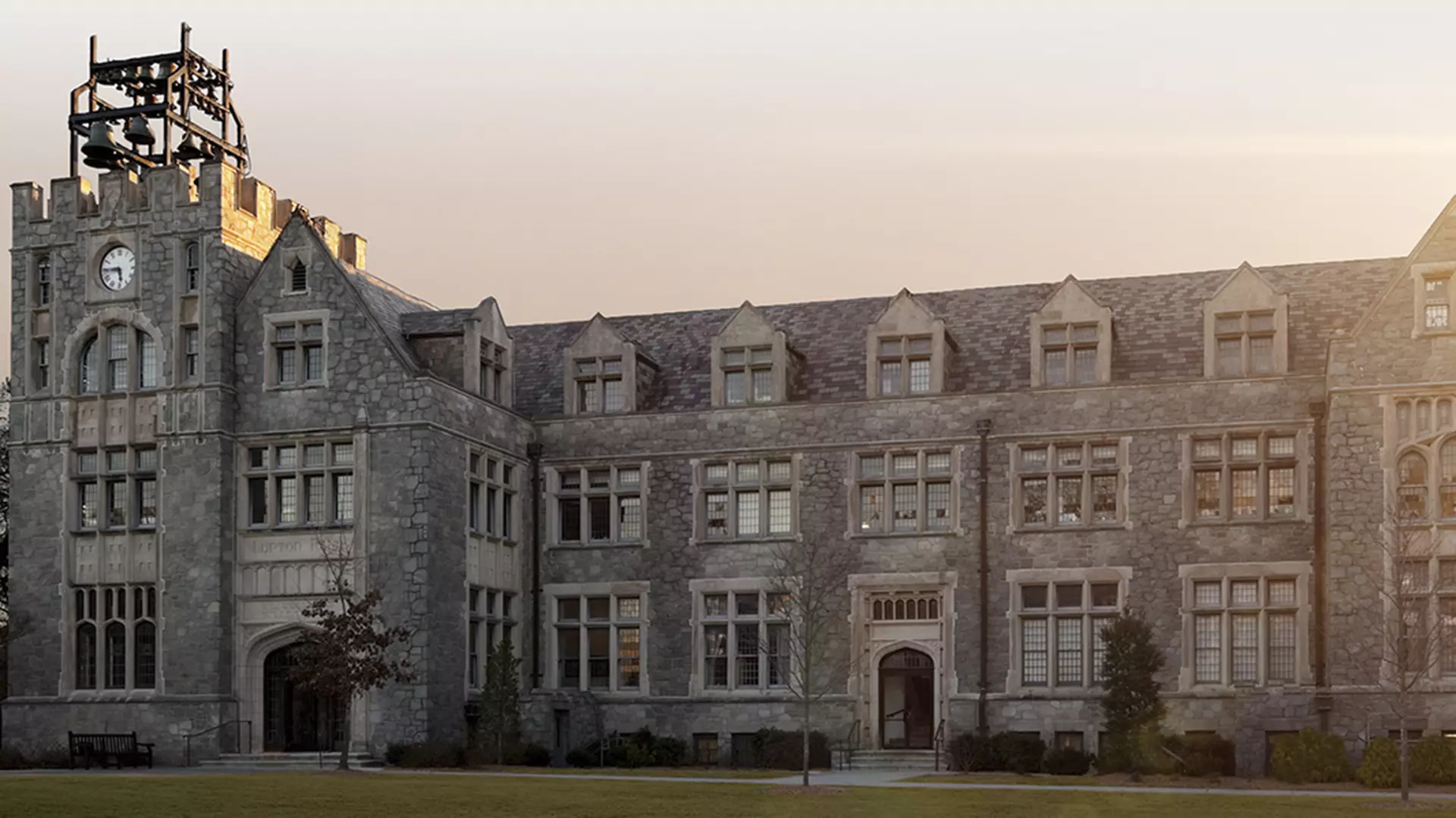 Historic stone university building with a clock tower and bell frame on the left at sunset over a lawn.