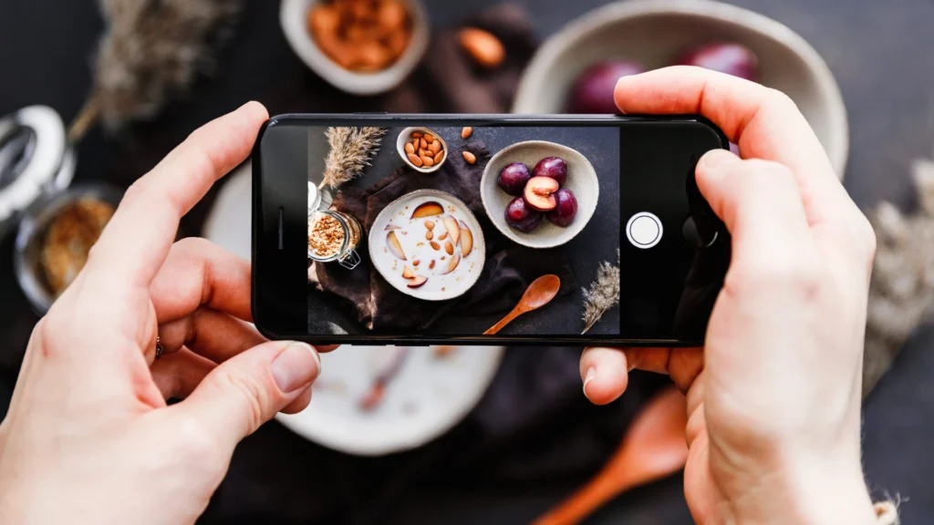 Social Media Video Phone Food Photography - See It Media Hands holding a smartphone photographing a styled bowl of yogurt, plums and almonds on a dark surface